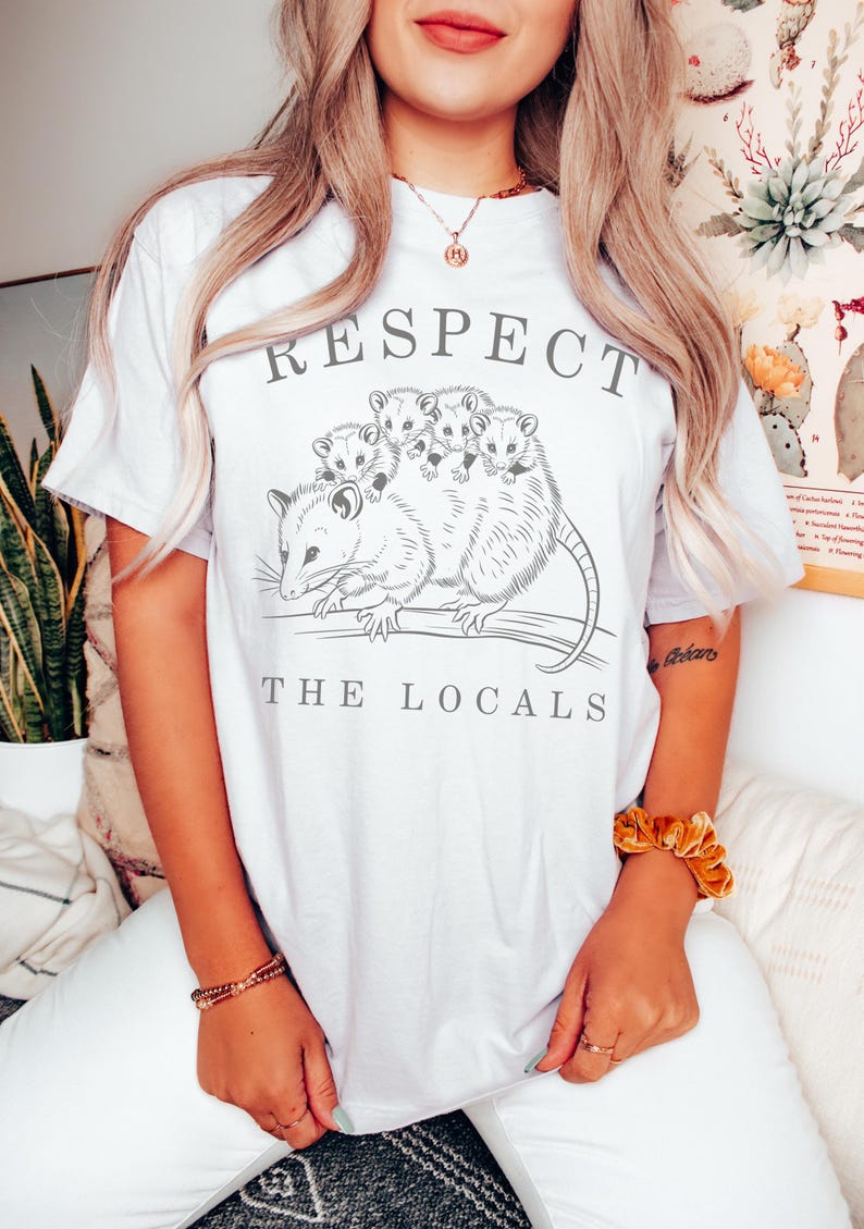 Puede incluir: Camiseta blanca con un gr&aacute;fico de una zarig&uuml;eya y sus cr&iacute;as, con las palabras "RESPECT THE LOCALS" en letras grises. La ilustraci&oacute;n de la zarig&uuml;eya es detallada y el texto est&aacute; en una fuente sencilla. La camiseta es de estilo casual.