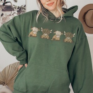 May include: A dark green hoodie with a design of four cartoon Highland cows. The cows are in shades of brown and cream, with fluffy fur and small horns. The hoodie has a drawstring hood and a front pocket, typical of a casual sweatshirt.