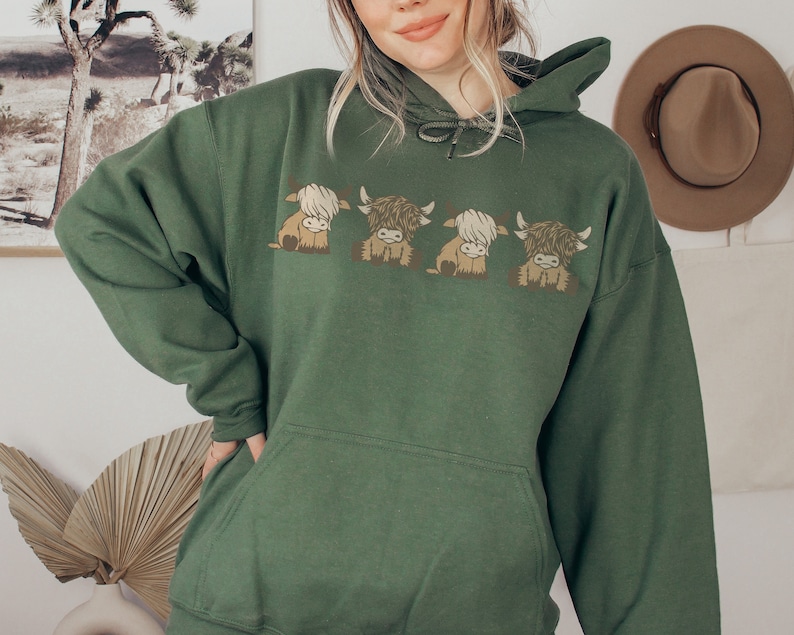 May include: A dark green hoodie with a design of four cartoon Highland cows. The cows are in shades of brown and cream, with fluffy fur and small horns. The hoodie has a drawstring hood and a front pocket, typical of a casual sweatshirt.