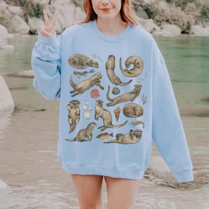Peut inclure: Sweat-shirt bleu clair orn&eacute; d'illustrations d'otaries dans diverses poses, accompagn&eacute;es de poissons et de coquillages. Les loutres sont de tons bruns et beiges, cr&eacute;ant un motif sur le th&egrave;me de la nature.