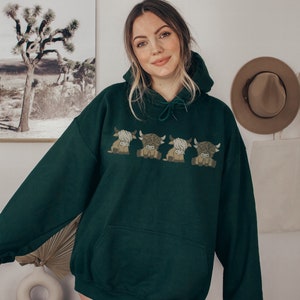 May include: A dark green hoodie with an embroidered design of four cartoon Highland cows. The hoodie has a drawstring hood and a kangaroo pocket. The cows are in shades of brown and tan.