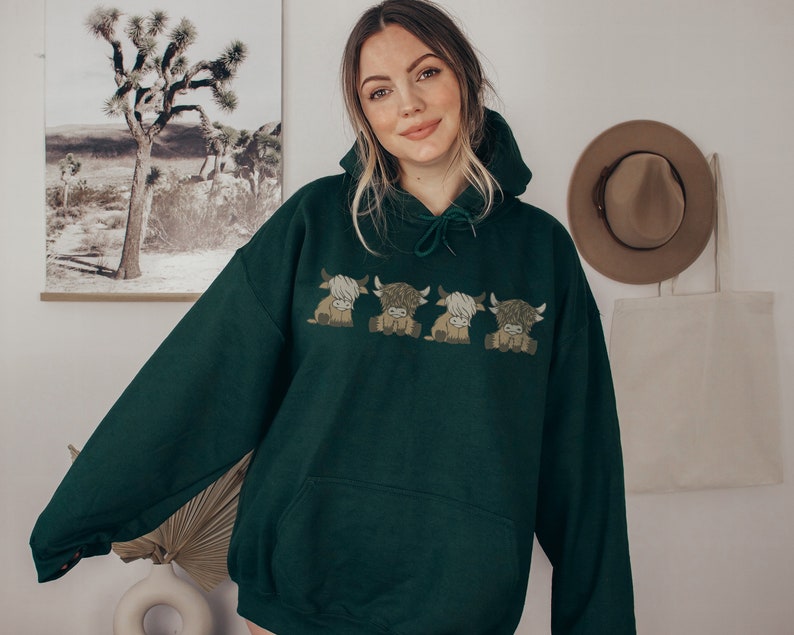 May include: A dark green hoodie with an embroidered design of four cartoon Highland cows. The hoodie has a drawstring hood and a kangaroo pocket. The cows are in shades of brown and tan.