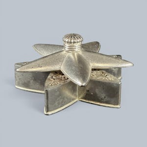 May include: A star-shaped silver-toned metal trinket box with a removable lid. The lid features a central ribbed knob and a smooth, pointed star design. The base of the box has intricate, cut-out floral patterns along the top edges of each point.