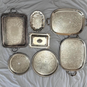May include: A collection of seven silver-plated serving trays and platters in various shapes, including rectangular, oval, and circular designs. Each piece features intricate embossed floral and scrollwork patterns along the borders and centres.