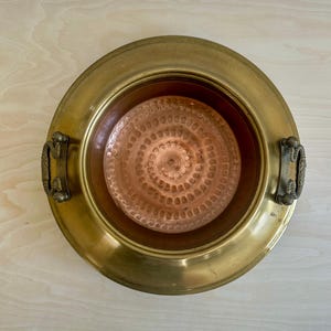 May include: A round brass bowl featuring a wide, smooth rim with fine concentric line detailing. The interior base is made of copper and displays a distinct hammered, circular pattern. Two ornate, textured brass handles are attached to the sides.
