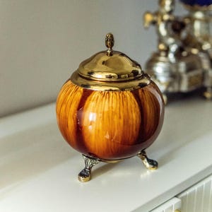 May include: A round ceramic jar with a glossy amber and brown drip glaze finish. The jar features a polished gold-tone metal lid with a small finial knob and rests on three ornate, textured gold-tone metal feet that elevate the vessel.