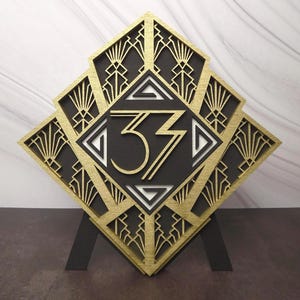 Peut inclure: Panneau en bois de style Art Déco, doré et noir, avec le chiffre "33" au centre. Le panneau en forme de losange présente des motifs géométriques complexes et est soutenu par un support noir.