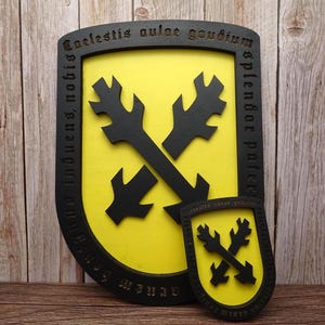 Peut inclure: Décoration murale en forme de bouclier noir et jaune avec un motif de croix. Le bouclier a une bordure noire avec du texte gravé. Une version plus petite du bouclier est placée devant le plus grand. Le fond est une surface en bois.