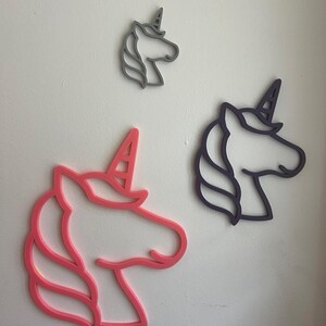 May include: Three unicorn head wall decorations in pink, grey, and purple. The unicorn heads are outlined with a horn and mane. The largest is pink, the medium is purple, and the smallest is grey. They are mounted on a white wall.