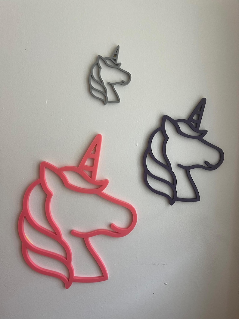 May include: Three unicorn head wall decorations in pink, grey, and purple. The unicorn heads are outlined with a horn and mane. The largest is pink, the medium is purple, and the smallest is grey. They are mounted on a white wall.