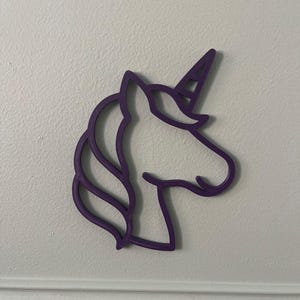 May include: A purple unicorn head silhouette, with a horn, mane, and facial details. The outline is a solid, vibrant purple, creating a whimsical and decorative element. The unicorn is mounted on a white wall.