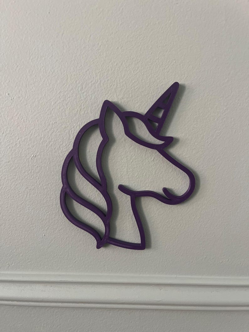 May include: A purple unicorn head silhouette, with a horn, mane, and facial details. The outline is a solid, vibrant purple, creating a whimsical and decorative element. The unicorn is mounted on a white wall.