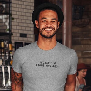 May include: A man wearing a grey t-shirt with the text "I worship a stone roller." printed on the front. He is smiling and wearing a black hat.