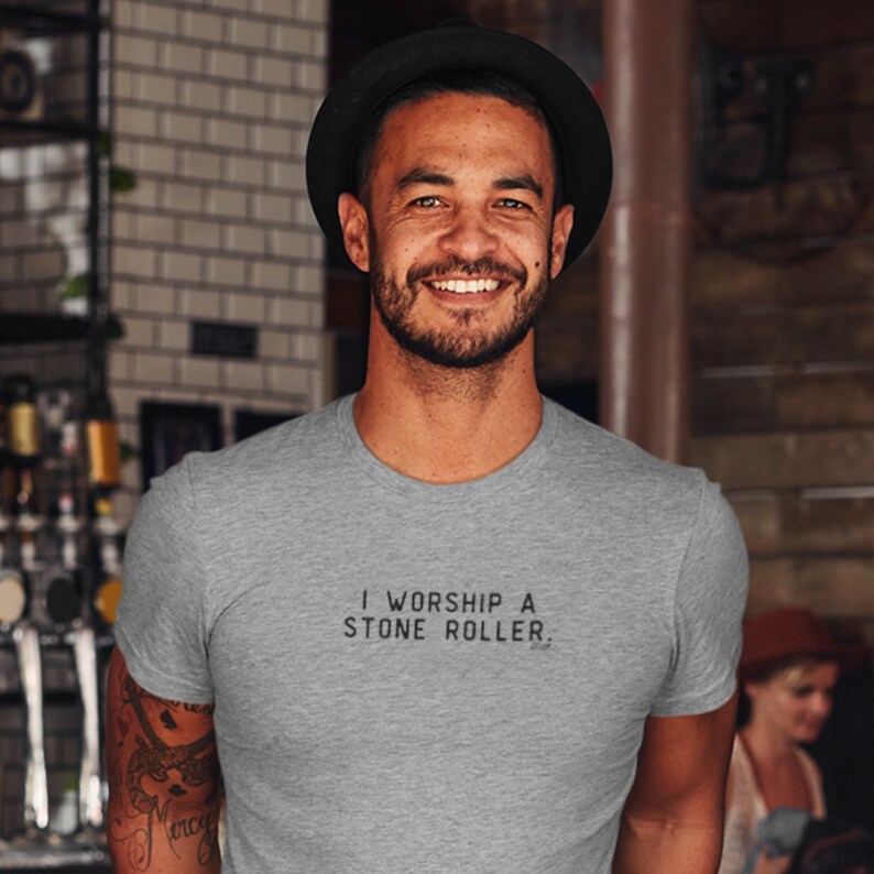 May include: A man wearing a grey t-shirt with the text "I worship a stone roller." printed on the front. He is smiling and wearing a black hat.