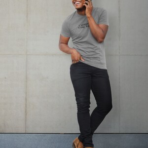 May include: A man wearing a heather grey t-shirt with the text "I Worship Stone Cold" printed on the front. He is wearing black jeans and brown shoes. He is standing in front of a concrete wall.