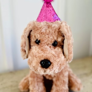 May include: A brown plush dog wearing a pink glittery party hat with a pink pom-pom on top.