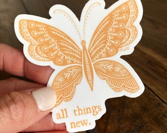 May include: A white sticker featuring an orange butterfly design with intricate patterns. The butterfly has detailed wings and a central body. The text "all things new" is printed in orange below the butterfly. The sticker has a decorative aesthetic.