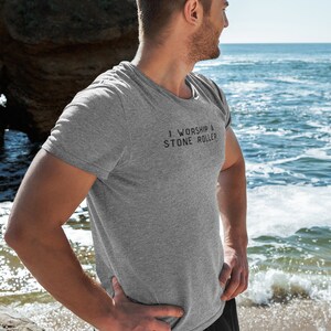 May include: A grey t-shirt with the text "I worship a stone roller" printed on the front. The person wearing the shirt is standing on a beach.