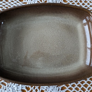 May include: Rectangular ceramic serving dish with rounded corners. The dish has a gradient colour scheme, transitioning from a dark brown rim to a lighter tan centre. The surface has a speckled texture and a glossy finish, suitable for serving food.