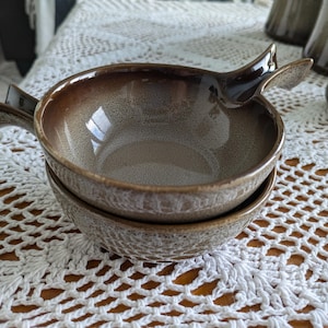 May include: Two brown ceramic bowls stacked, featuring handles. The bowls have a speckled, glazed finish and a textured exterior. The handles are shaped like bird wings. Other matching bowls are visible in the background.
