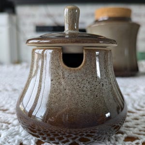 May include: A brown ceramic sugar bowl with a lid and a small handle. The bowl has a speckled pattern and a glossy finish. The lid has a small handle. Other matching ceramic jars are in the background.