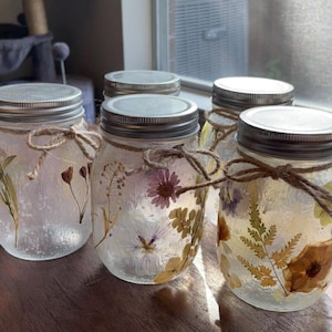 May include: Five clear glass jars with silver lids, filled with dried flowers and greenery. The jars are arranged in a row on a wooden surface.