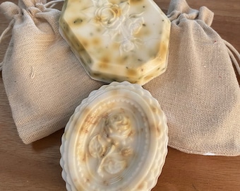 May include: Two handmade soap bars with a floral design. The soaps are in burlap bags. One soap is a hexagonal shape and the other is oval.