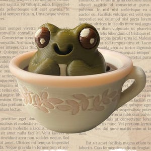 Könnte beinhalten: Eine 3D-gedruckte Froschfigur sitzt in einer dekorativen Teetasse. Der Frosch ist grün mit braunen und weißen Augen, und die Teetasse ist beige mit einem rosa Rand und floralem Design. Die Teetasse steht auf einem Hintergrund aus Vintage-Papier.