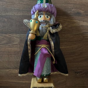 May include: A decorative nutcracker figurine of a king, wearing a green and purple turban and a black cape with gold trim. The king holds a gold vessel and stands on a wooden base. The base has text that reads "C+M+B Melchior C+M+B".