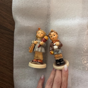 May include: Two vintage porcelain figurines of children. The girl wears a gray dress, holds a school cone, and carries a brown satchel. The boy wears a brown jacket, a scarf, and holds an apple. Both stand on round bases.