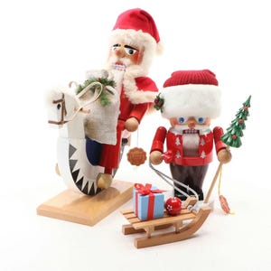 May include: Three Christmas-themed wooden nutcrackers. One is Santa Claus riding a white horse with a sack of gifts. Another is a nutcracker in a red jacket holding a Christmas tree. A small wooden sled with a gift box and ornament is in front.