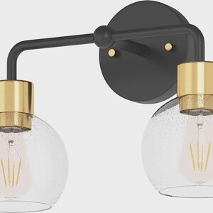 May include: A two-light wall sconce featuring a matte black circular backplate with two small gold-toned fasteners. The fixture has a black horizontal arm supporting two downward-facing clear glass globes with seeded texture and gold-toned sockets.