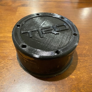 May include: Black plastic center cap for a car wheel with the word "TRD" embossed on the top.