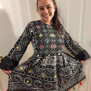 May include: A black dress with a white and colorful geometric pattern. The dress has a round neckline, long sleeves, and a flared skirt. The dress is decorated with small mirrors and tassels.
