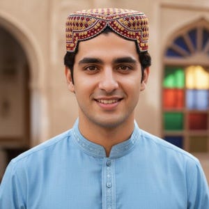 May include: A man wearing a traditional embroidered cap with a red and gold pattern. He is wearing a light blue tunic with a mandarin collar. The background features an arched doorway and a stained-glass window.