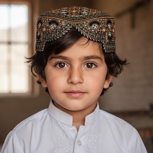 May include: A young person wearing a traditional embroidered cap with a detailed pattern in shades of brown, gold, and black. The person is also wearing a white embroidered shirt. The background is a neutral color.