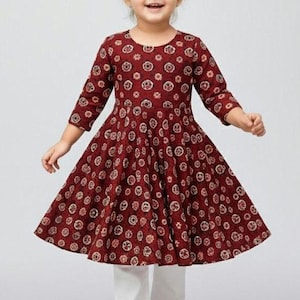 May include: A young girl wearing a maroon dress with a floral pattern and white pants. The dress has three-quarter sleeves and a flared skirt. The girl is also wearing white sandals. Her hair is styled in two buns.