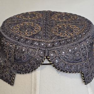 May include: A dark gray cap with intricate gold embroidery. The cap features a rounded crown and a scalloped brim, with detailed patterns and embellishments. The design includes floral and geometric motifs, creating a decorative headwear piece.