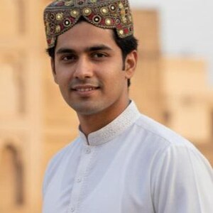 May include: A man wearing a traditional cap with intricate embroidery in gold, red, and green. The cap is paired with a white collared shirt. The background is a blurred outdoor setting.