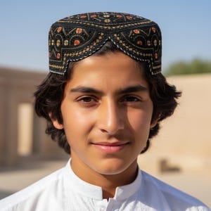May include: A black embroidered cap with a geometric pattern, featuring red and gold accents. The cap is worn with a white shirt. The image is a portrait of a person wearing the cap.