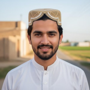 May include: A man wearing a beige and white crocheted cap with a geometric design. He is also wearing a white collared shirt. The background shows a road and buildings.