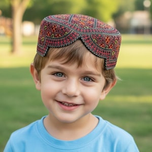 May include: A child wearing a red, black, and yellow embroidered cap. The cap has a curved design along the bottom edge and a detailed pattern. The child is wearing a light blue shirt and smiling.