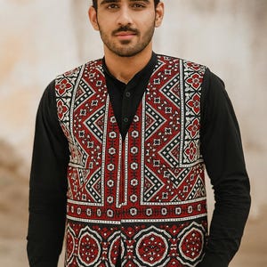 Op de afbeelding: Een vest met een rood, zwart en wit patroon. Het vest wordt gedragen over een zwart shirt met lange mouwen. Het vest heeft geometrische en bloemmotieven. De persoon draagt een zwarte broek.