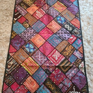 May include: A colorful patchwork wall tapestry with a diamond pattern. The tapestry is made of various fabrics and features embroidered details and mirrored accents.