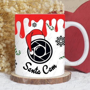 Puede incluir: Taza de cerámica blanca con un gorro de Papá Noel rojo y un diseño de lente de cámara negra. La taza tiene pintura roja goteando y el texto "Santa Cam" en la parte delantera.