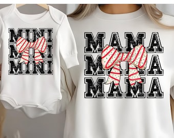 Op de afbeelding: Witte sweatshirt met lange mouwen en babyromper met bijpassende ontwerpen. De sweatshirt heeft het woord "MAMA" drie keer gestapeld en de romper heeft "MINI" drie keer gestapeld. Beide hebben een rood-witte strik-afbeelding.