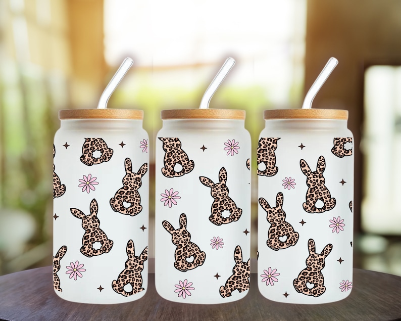May include: Three glass tumblers with bamboo lids and clear straws. Each tumbler features a white background with a repeating pattern of pink flowers and brown bunnies with leopard print fur.