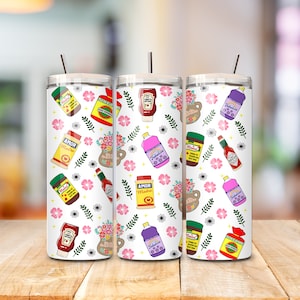 May include: Three white stainless steel tumblers with lids and straws. The tumblers are decorated with a pattern of pink flowers, green leaves, and various food and beverage items, including bottles of hot sauce, ketchup, and cleaning products. The text "Amor Madre" and "Fabulous" are visible on some of the items.