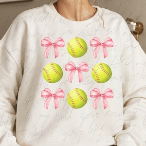May include: White sweatshirt with a pattern of pink bows and yellow softball graphics.