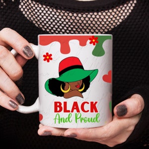 Puede incluir: Taza de cerámica blanca con un gráfico de una mujer negra con un sombrero verde con borde rojo y pendientes de aro dorados. La taza tiene las palabras "BLACK And Proud" en rojo y verde.